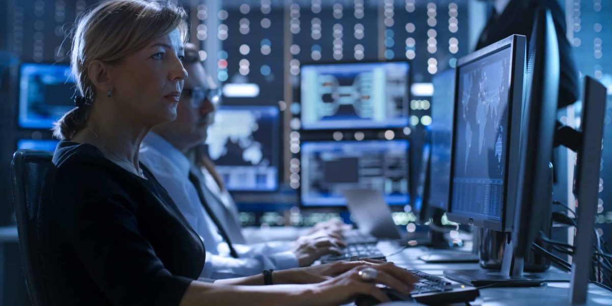 Female Government Employee Works in a Monitoring Room. In The Background Supervisor Holds Briefing. Possibly Government Agency Conducts Investigation.