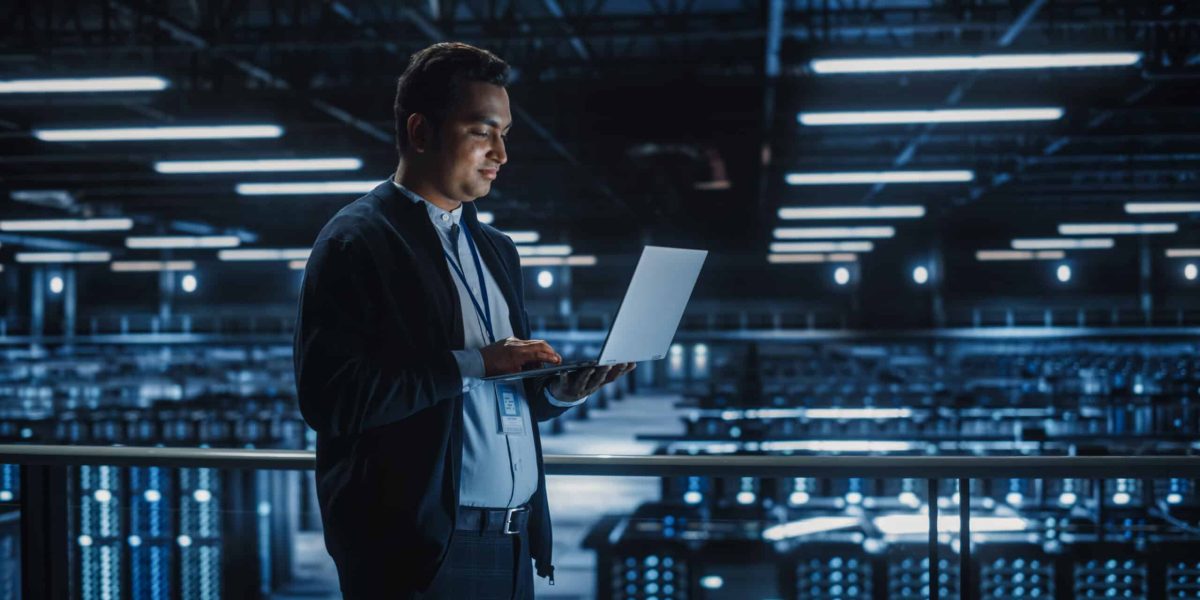 Portrait of IT Specialist Uses Laptop in Data Center. Server Farm Cloud Computing Facility with Male System Administrator Working. Cyber Security and Network Protection.